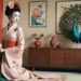 A woman in a pink kimono with floral patterns sits next to a peacock, with another peacock standing on a wooden cabinet in a traditional Japanese-style room.