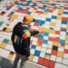 Person in yellow cap and colorful jacket pointing at large floor calendar with multicolored squares and numbers.